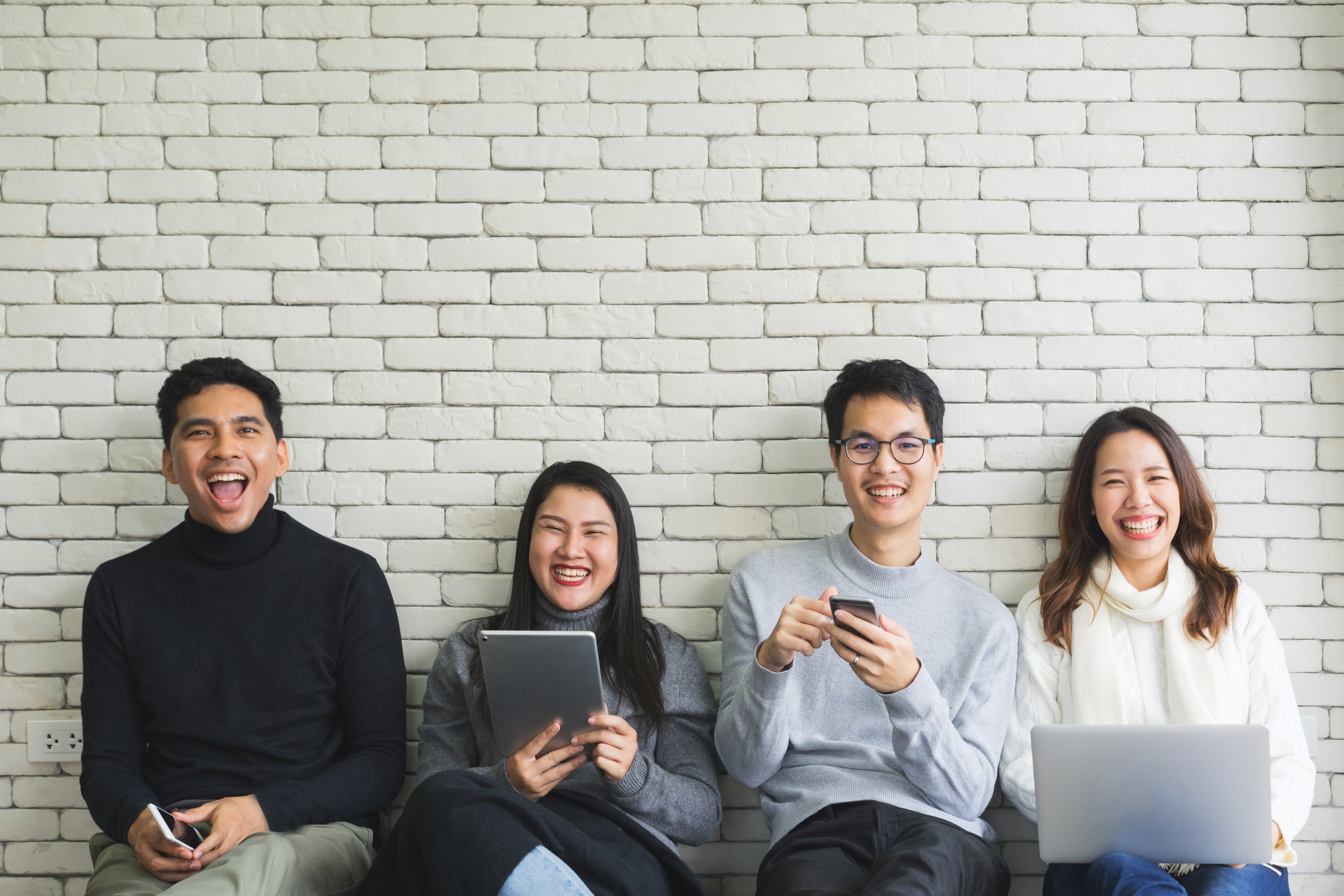 Image of close up group of millennial generation teenagers holding modern gadget devices and sitting on white brick background wall room, technology with lifestyle concept